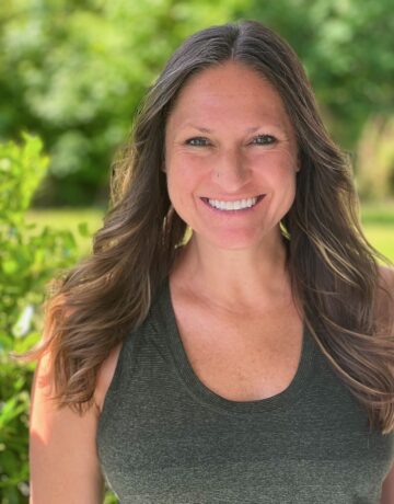 A woman wearing a green tank top smiles for the camera. She is surrounded by Green plants.