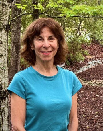 A woman in a bright blue t-shirt stands near a tree.