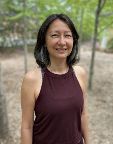 A woman standing in the woods wearing a maroon shirt.