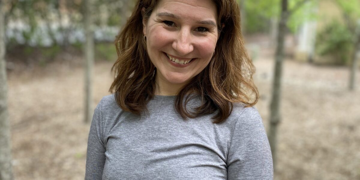 A woman standing in the woods wearing a gray shirt.