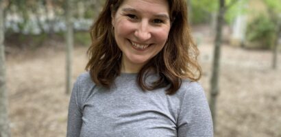 A woman standing in the woods wearing a gray shirt.