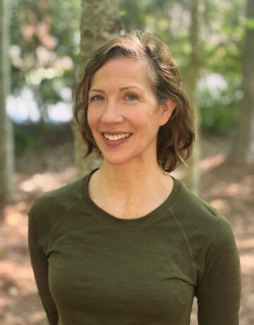 A woman with short wavy hair smiles. She wears a green long sleeve shirt and stands in the woods.