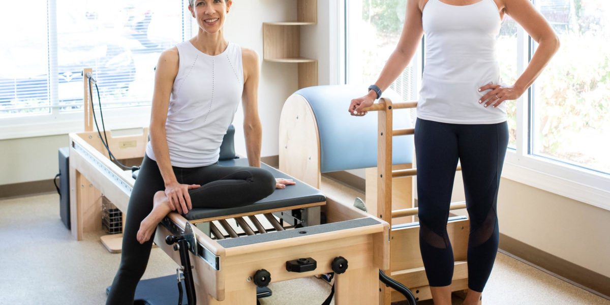 Two women standing next to a pilates machine.