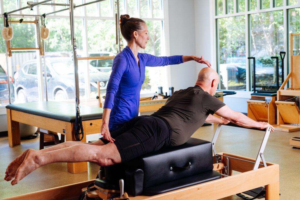 Deborah instructs a client on the Pilates Reformer. Her hand makes contact with the top of his head.