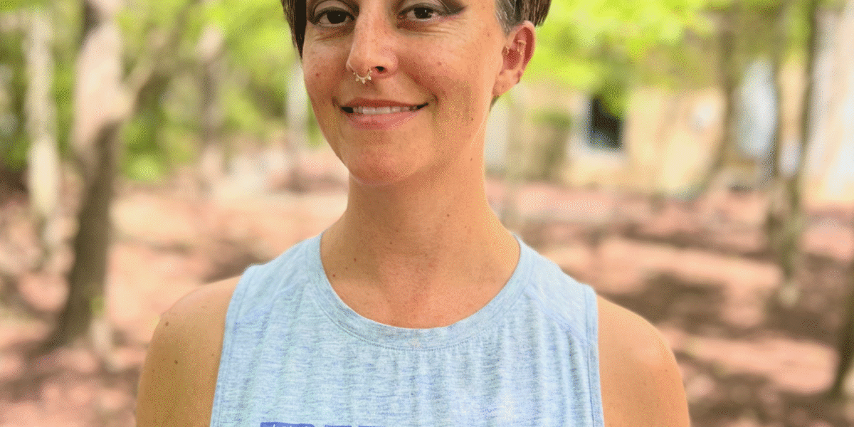 A woman with short hair wears a grey PUMA branded tank top. She smiles, surrounded by woods.