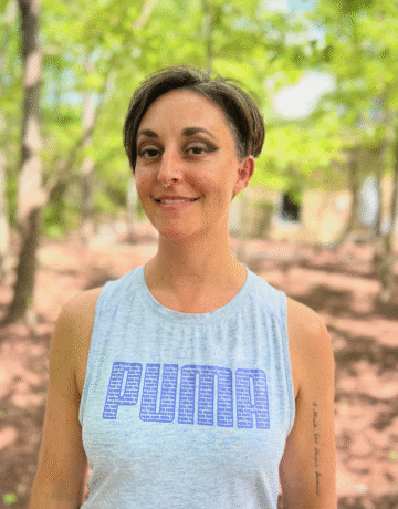 A woman with short hair wears a grey PUMA branded tank top. She smiles, surrounded by woods.