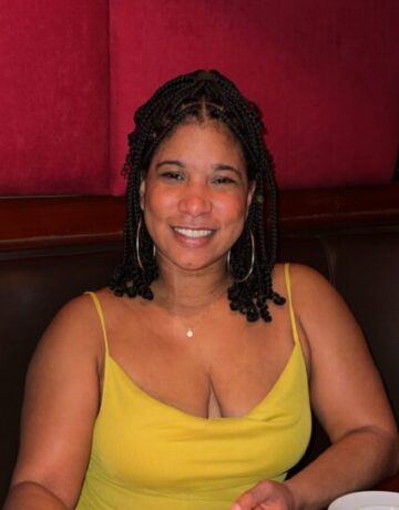 A woman wearing a yellow dress and hoop earrings smiles at the camera.